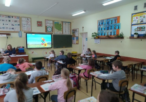 Uczniowie siedzą w ławkach.W tle widoczne ściany klasy z pomocami dydaktycznymi oraz tablica interaktywna.